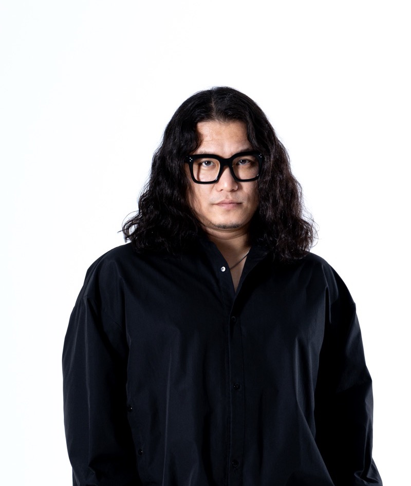 白い背景の前で、黒いシャツを着て正面を向く華僑J。メガネをかけている。