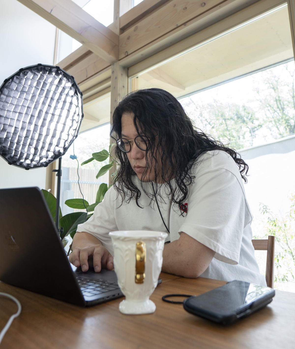 パソコンに向かって作業をする華僑J。メガネをかけ、窓の外の光が入る部屋にいる。